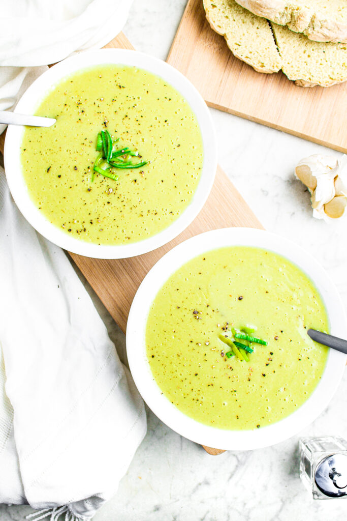 Overhead view of two round white bowls with creamy vegan potato leek soup topped with chopped leeks and ground black pepper flakes