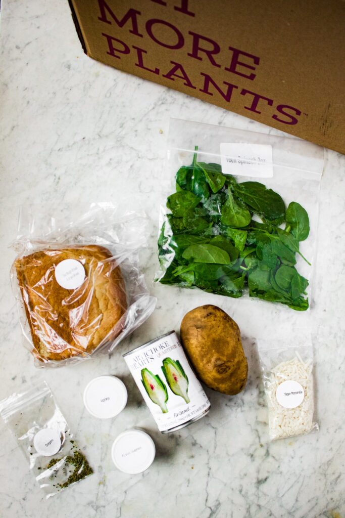 Overhead shot of all the ingredients from a Purple Carrot meal delivery box to make Vegan Spinach Artichoke Grilled Cheese: a bag of spinach, potato, canned artichoke hearts, baggie of vegan mozzarella, vegenaise, a bag of sourdough bread slices, oregano, and pickled jalapenos.