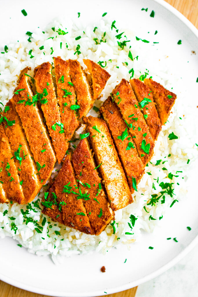 Overhead view of a round white plate with a vegan curry tofu cutlet sliced diagonally on a bed of white rice sprinkled with parsley