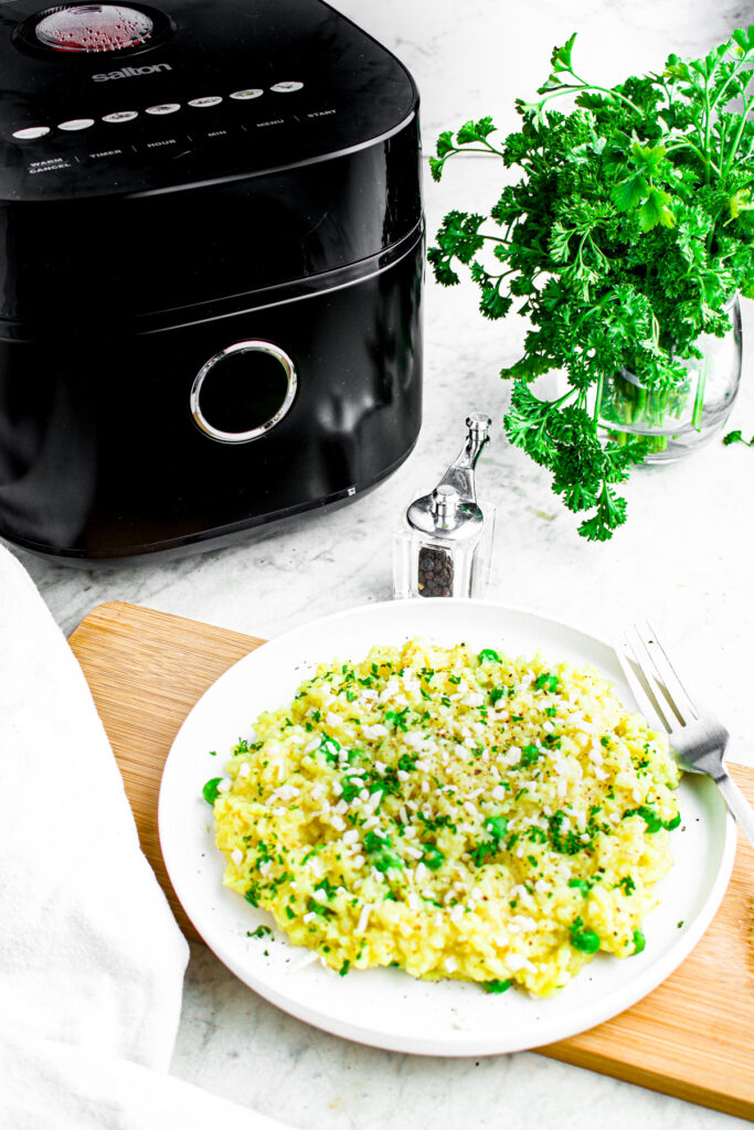 Overhead view of a round white plate of vegan risotto in front of a sleek black Salton Micom rice cooker