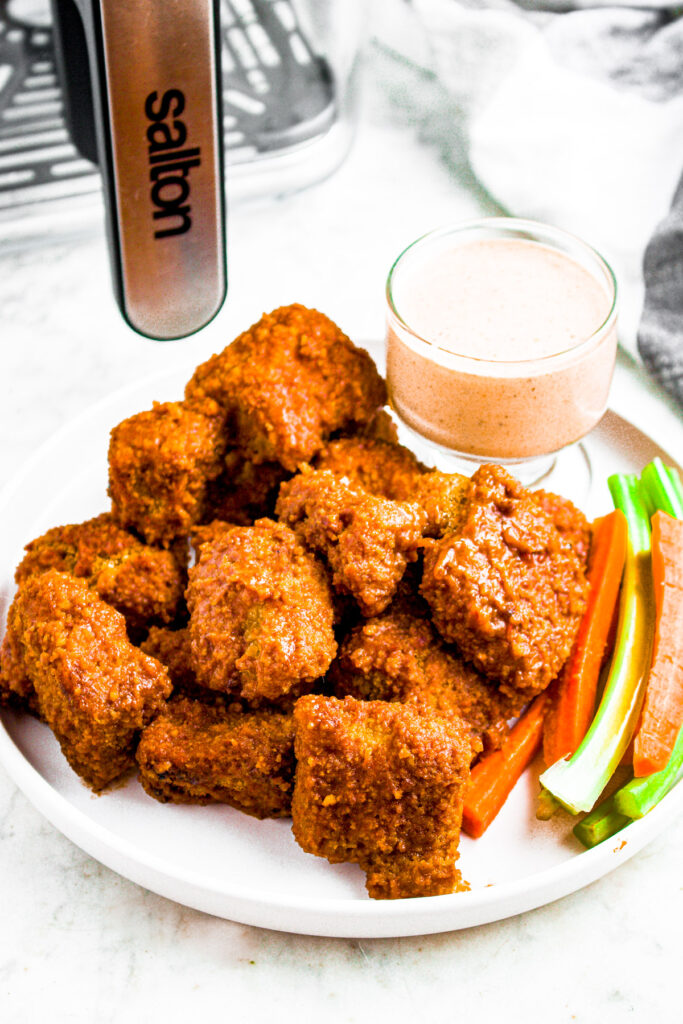 Overhead photo of a pile of bbq vegan wings on a round white plate next to a glass bowl of vegan bbq ranch and some celery and carrot sticks