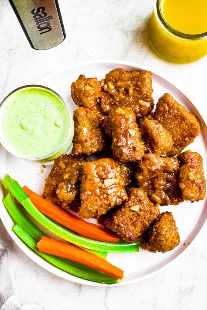 Overhead photo of a round white plate piled high with pineapple tofu wings with celery sticks and carrots and a small bowl of avocado ranch