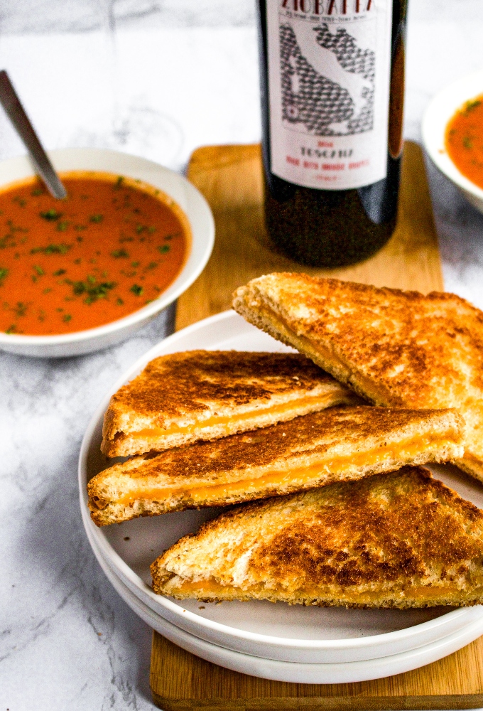 A plate with a pile of vegan grilled cheese sandwiches cut in half to make triangles. There is a bowl of dairy-free cream of tomato soup and a bottle of sangiovese in the background.