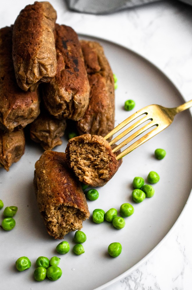Head on photo of a pile of vegan sausages on a round plate with green peas. One of the sausages is cut in half and has a fork sticking into it.