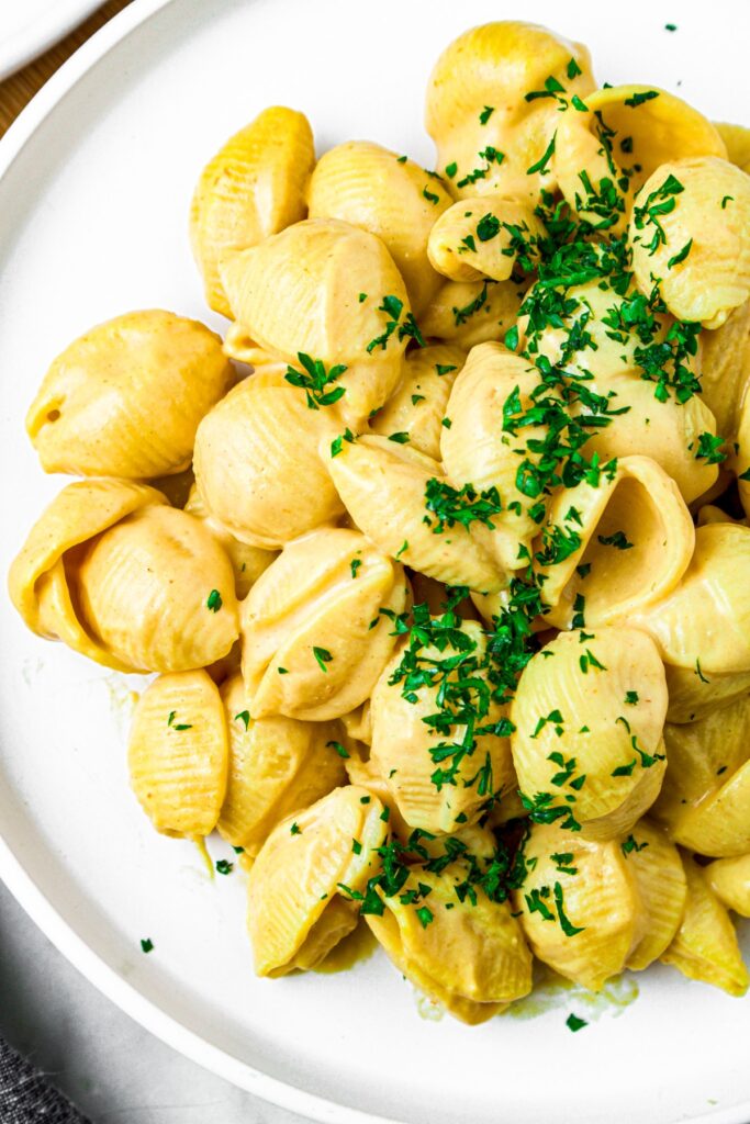 Overhead close up shot of a round white plate with a pile of vegan shells and cheese sprinkled with bright green herbs