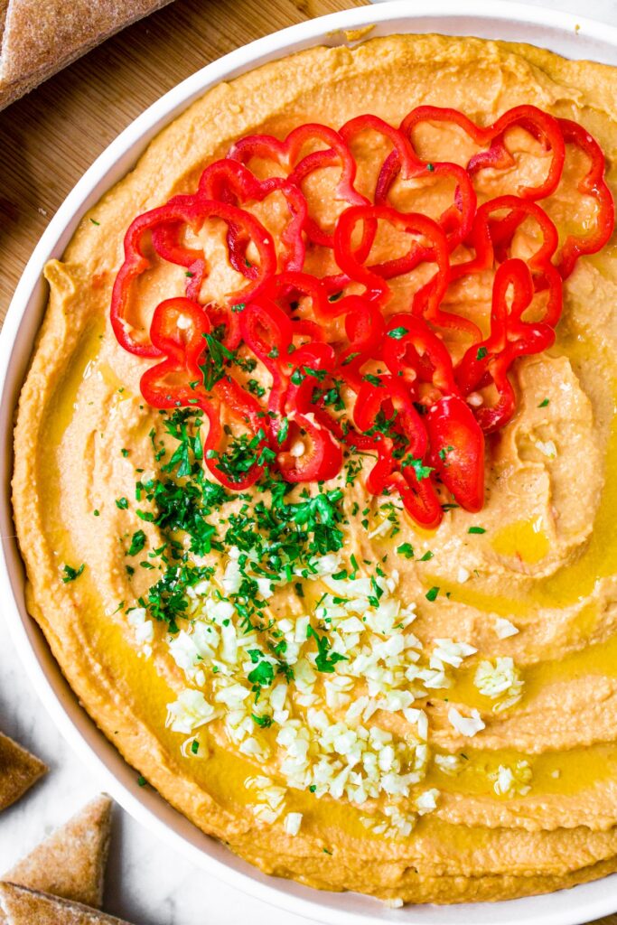 Overhead close up photo of a plate of spicy garlic hummus presented in a swirl pattern with olive oil drizzled into the crevices and topped with minced garlic, parsley, and sliced habanero peppers