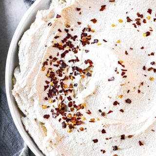 Close up overhead shot of a circular schmear of dairy-free ricotta on a round white plate sprinkled with red pepper flakes and surrounded by a gray linen