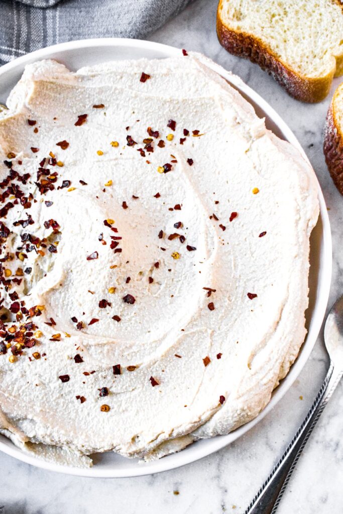 Overhead shot of a round white plate with a circular schmear of vegan ricotta cheese sprinkled with crushed red pepper flakes. There are small slices of bread in the top right corner.