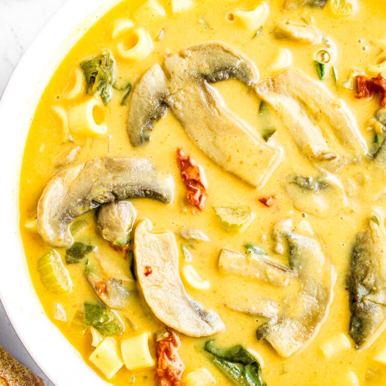Overhead close up photo of half a round white bowl filled with creamy Tuscan mushroom soup with sun-dried tomatoes, ditalini, and spinach