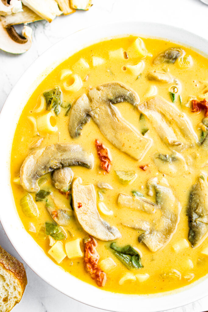 Overhead close up view of a round white bowl of creamy Tuscan vegetable soup with big meaty mushroom slices, sun-dried tomatoes, pasta, and spinach