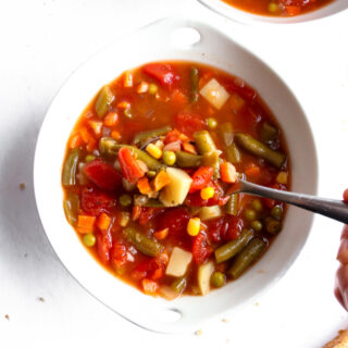 Overhead photo of homestyle vegetable soup in a round white bowl with a spoon digging into it and bread slices off to the side.