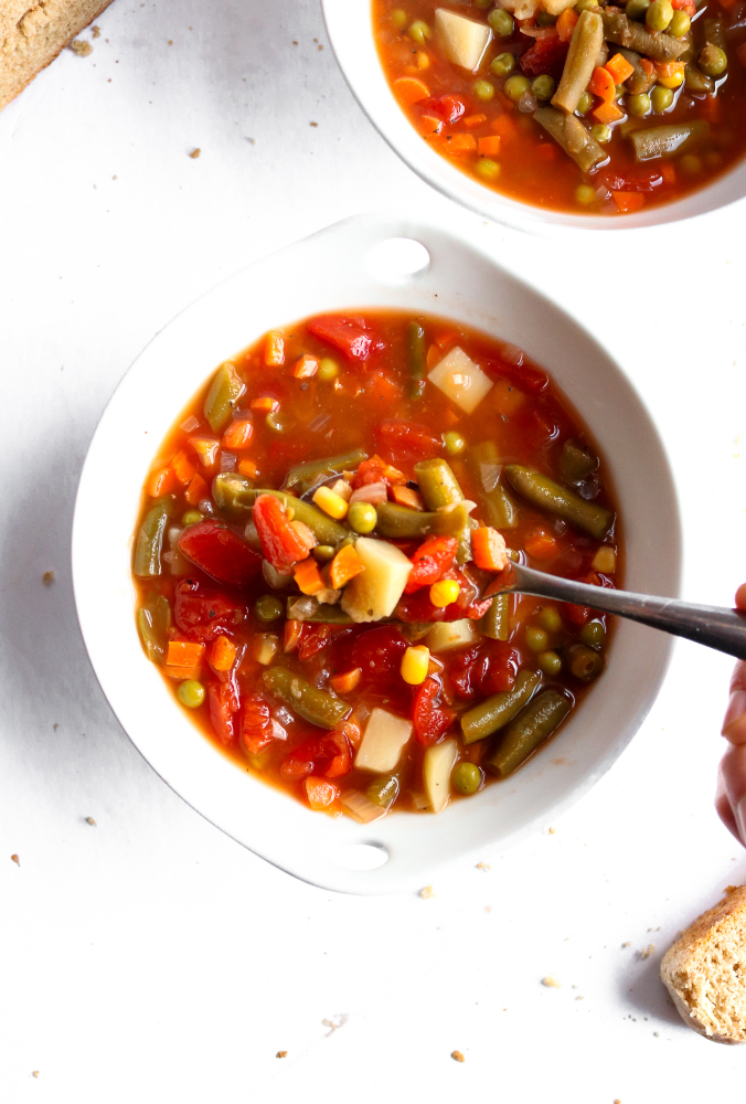 Overhead photo of homestyle vegetable soup in a round white bowl with a spoon digging into it and bread slices off to the side.