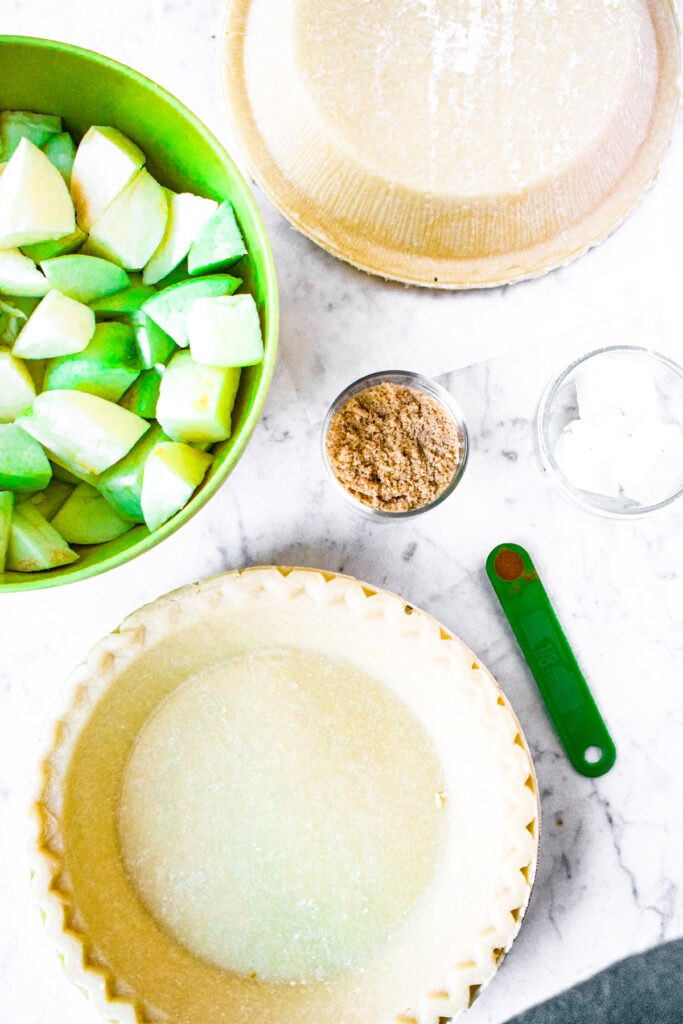 Overhead photo of all the ingredients you need to make dairy free apple pie: pie crust, apples, brown sugar, vegan butter, and cinnamon