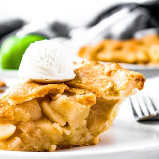 Close up head on photo of a slice of apple pie on a plate with vanilla ice cream on top and a green apple in the background