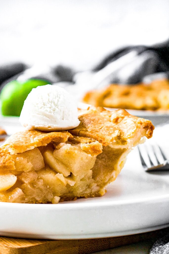 Close up head on photo of a slice of apple pie on a plate with vanilla ice cream on top and a green apple in the background