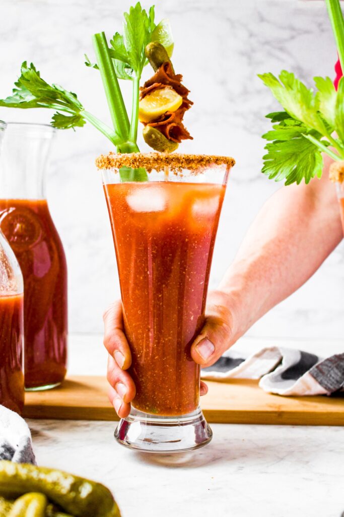 Head on photo of a hand picking up a tall glass of spicy Bloody Mary cocktail garnished with a stalk of celery