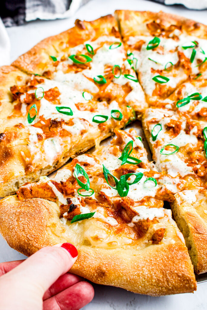 Overhead view of a vegetarian buffalo chicken pizza with a hand with red nail polish pulling one of the pieces out