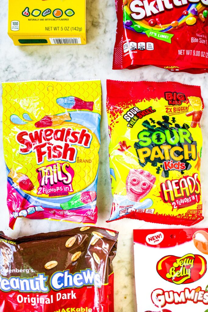 Overhead shot of 6 bags of various vegan-friendly candies: Chuckles, Skittles, Swedish Fish, Sour Patch Kids, Goldenberg's Peanut Chews, and Jelly Belly