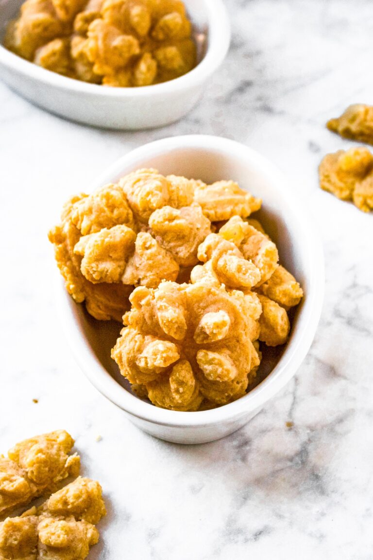 A head-on shot of flower-shaped vegan cheese straws arranged in a white oval shaped dish. There is a matching dish also filled with cheese straws in the background and more dairy free cheese straws around the two dishes.