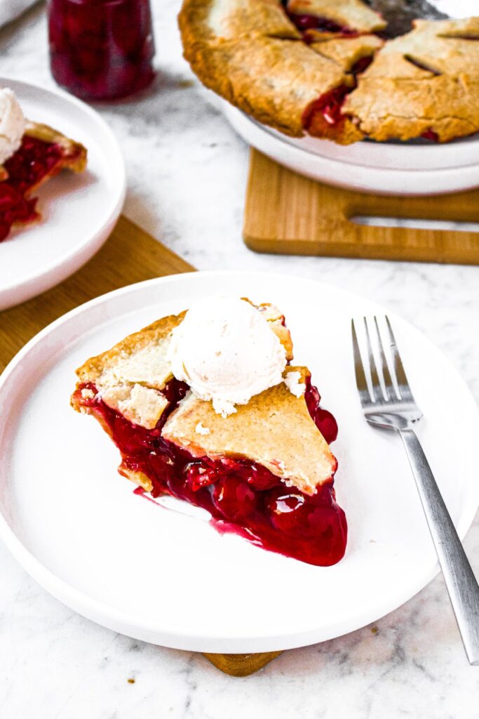 Overhead photo of a slice of vegan cherry pie on a plate next to a fork and topped with ice cream