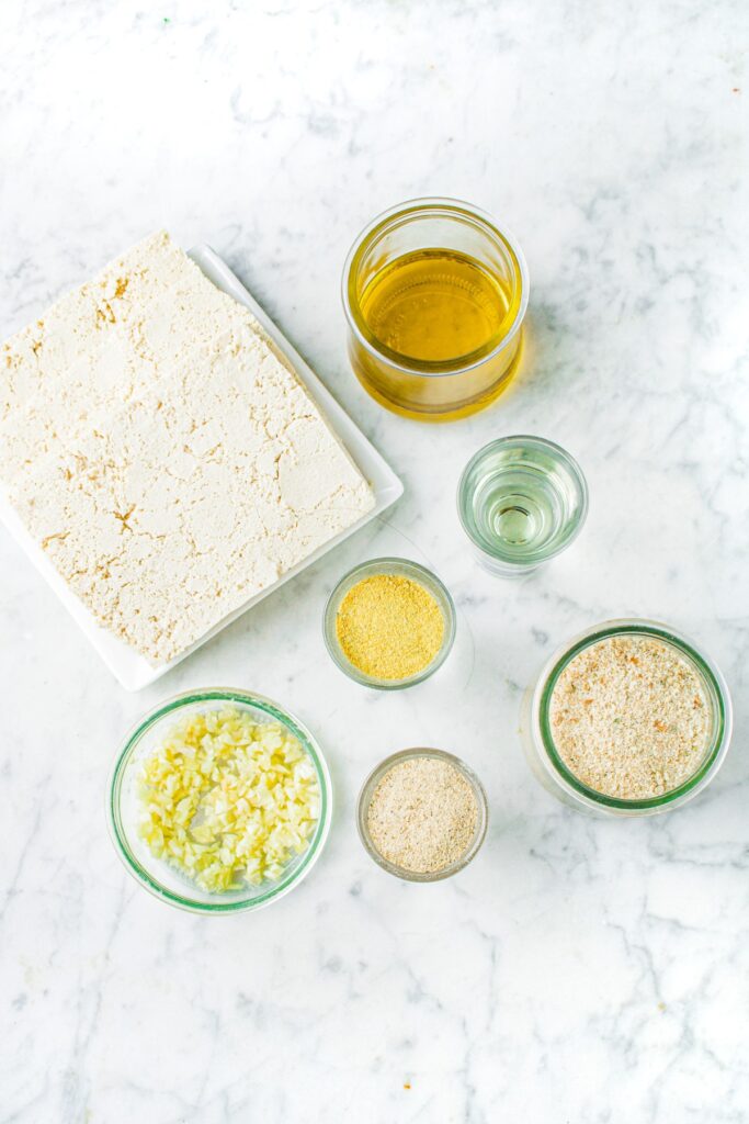 Overhead shot of the ingredients you need to make tofu schnitzel: sliced frozen and thawed tofu, olive oil, vinegar, vegan chicken bouillon powder, bread crumbs, garlic, and spices