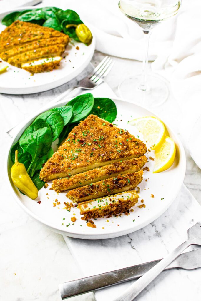 Overhead shot of a vegan chicken cutlet, partially sliced, on a plate with fresh spinach leaves, lemon wedges, and pepporincini. There's another identical plate behind the first plate along with two glasses of white wine.