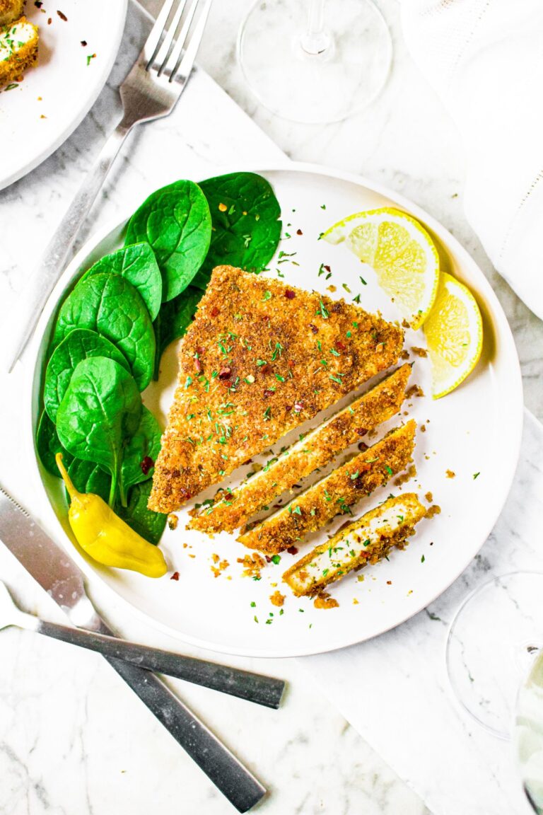 Overhead shot of a vegan chicken cutlet, partially sliced, on a plate with fresh spinach leaves, lemon wedges, and pepporincini.