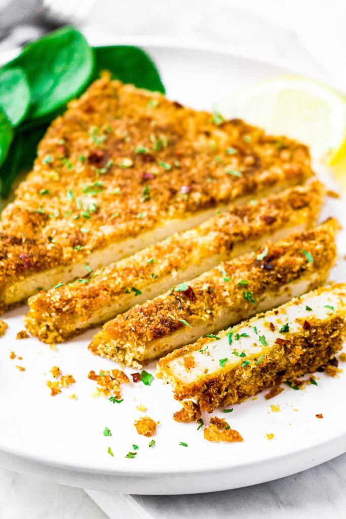 Close up shot of a sliced breaded tofu cutlet on a plate next to fresh spinach