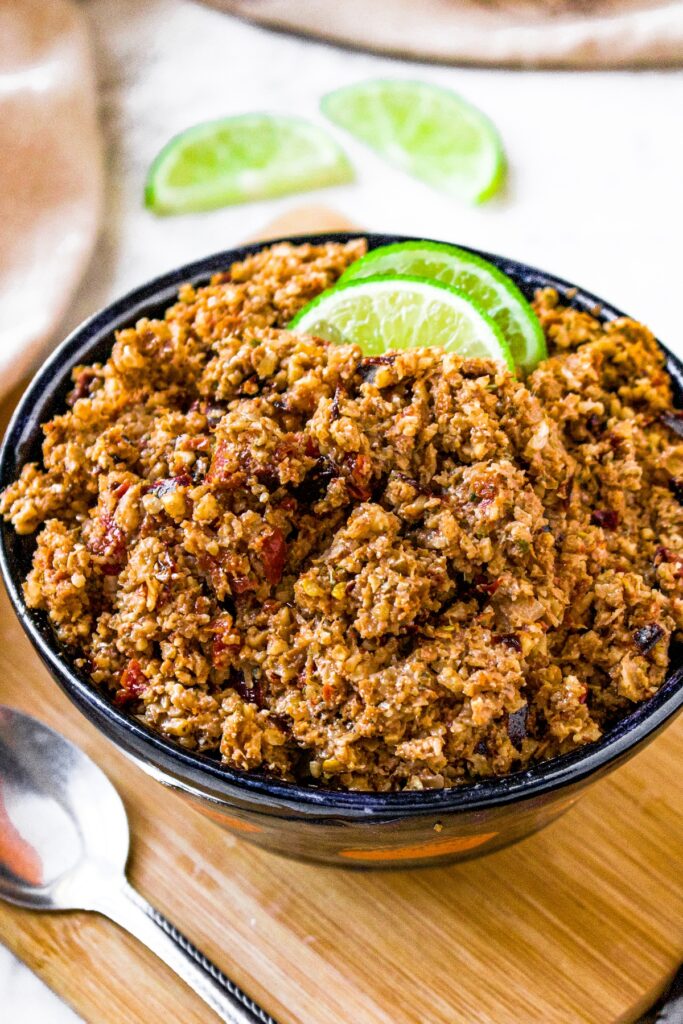 Close up overhead photo of a navy blue bowl filled with meatless walnut chorizo and 2 slices of lime. There are more lime slices behind the bowl and a spoon on the left side of the bowl.