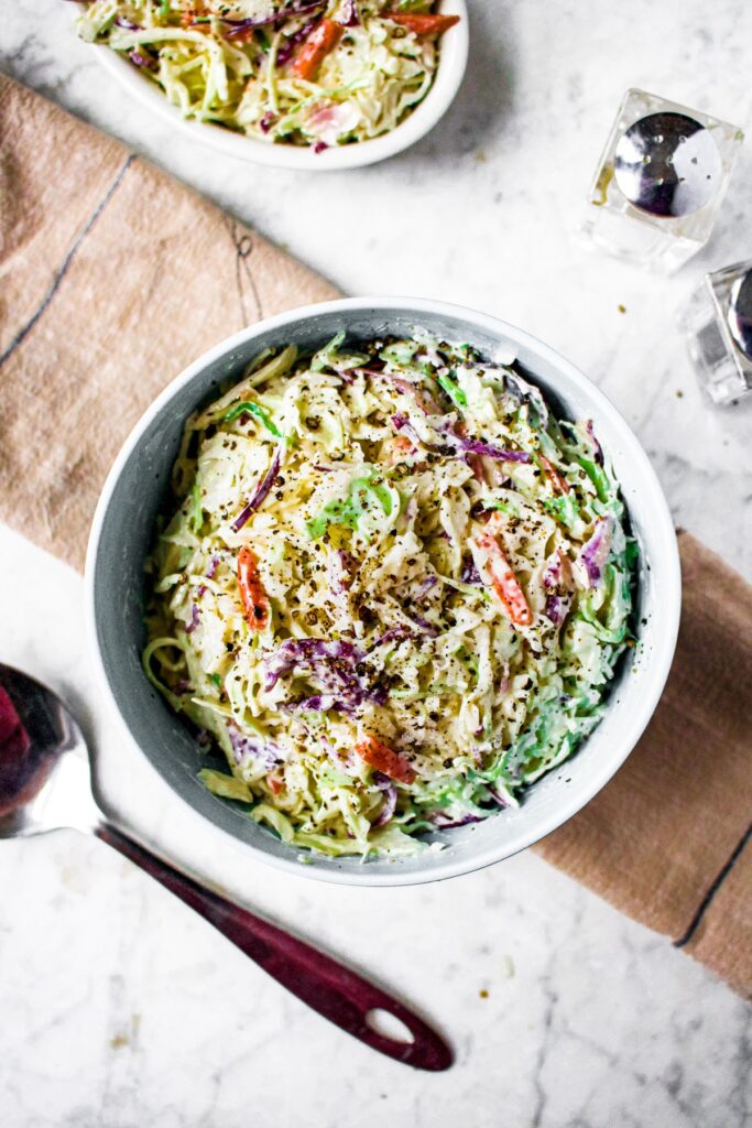 Overhead shot of a bowl of vegan coleslaw with a ramekin full of coleslaw to the side, a large metal spoon to the other side, salt and pepper shakers, and a folded beige linen