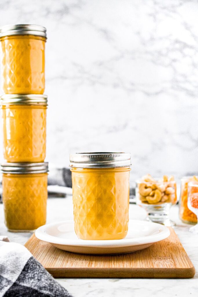 Head-on shot of a jar of cream of cheddar soup sitting on a small white plate. There is a tower of three more jars of soup to the left and just behind the front jar. There are clear cups, one filled with cashews and the other with red lentils, behind and to the right of the jar.