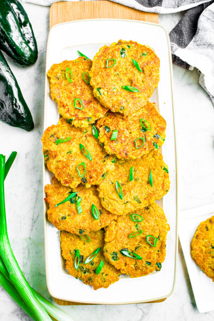 Overhead photo of a pile of vegan jalapeno corn cakes on a long white rectangular plate surrounded by peppers, green onions, and a black and white linen cloth