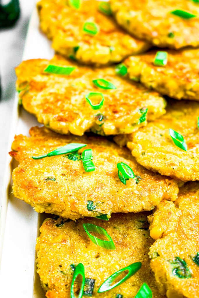 A pile of vegan corn fritters on a long white rectangular plate topped with sliced green onions