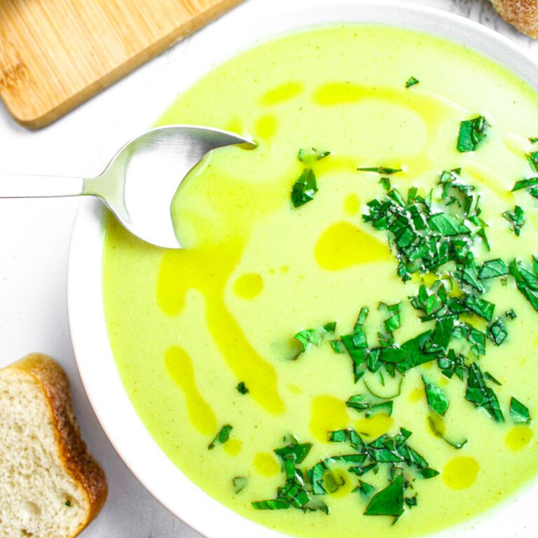 Overhead close up view of a silver spoon digging into a vegan cream of zucchini soup with olive oil swirl and fresh herbs