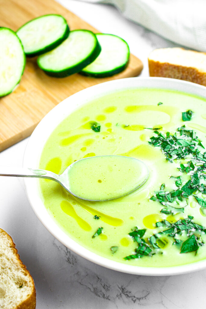 Overhead view of a spoon dipping into a round white bowl of vegan cream of zucchini with a half sliced zucchini and slice of french bread in the background