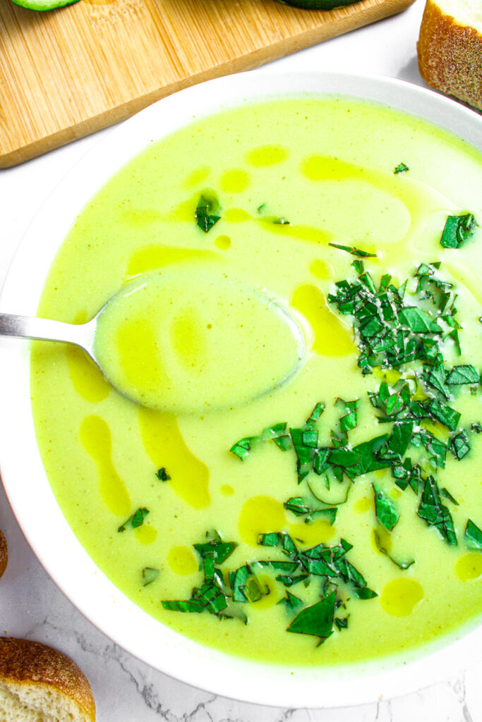 Overhead close up view of a round white bowl of creamy zucchini soup with a spoon coming out of it