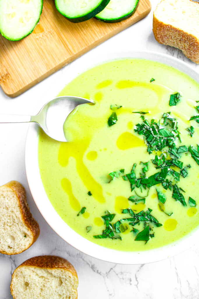 Overhead view of a smooth and creamy zucchini blender soup with a spoon digging into the left side and fresh basil garnish on the right side