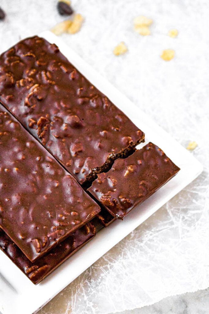 Overhead photo of two vegan crunch bars on a square white plate. One square of the chocolate bar is falling off the plate