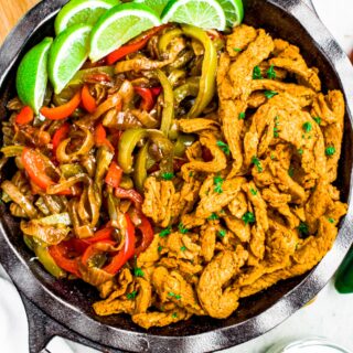 Overhead close up photo of a deep cast iron pan filled with vegan chicken, peppers and onions, and limes for fajitas