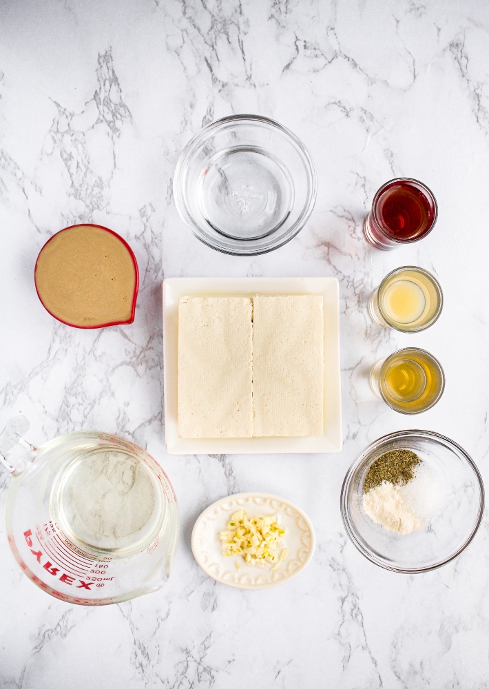 Overhead shot of the ingredients you need to make whipped vegan feta dip: firm tofu, refined coconut oil, lemon juice, kalamata brine, apple cider vinegar, tahini, water, minced garlic, and spices.