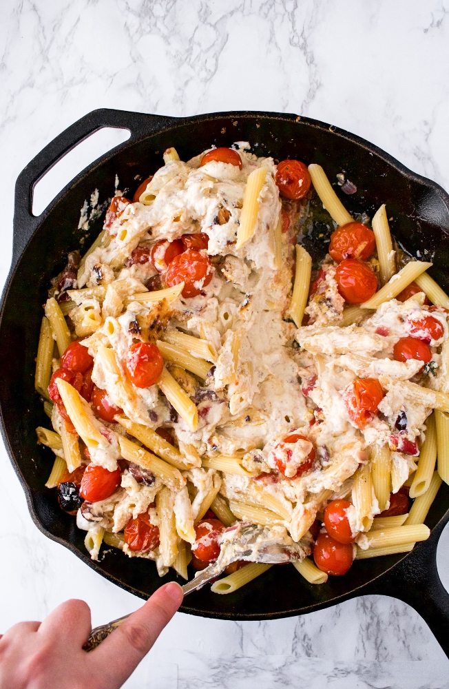 Overhead photo of a hand stirring pasta into a tofu feta sauce with cherry tomatoes in a cast iron pan
