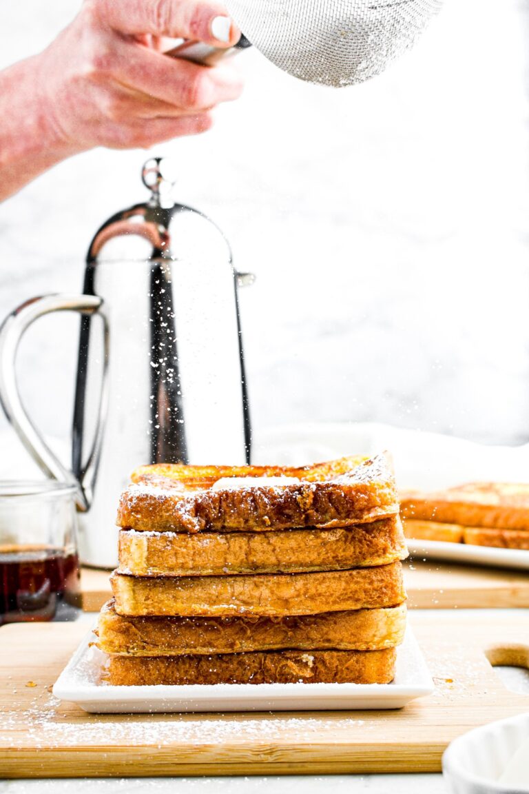 Head on photo of a stack of eggless French toast with a hand sifting powdered sugar on top