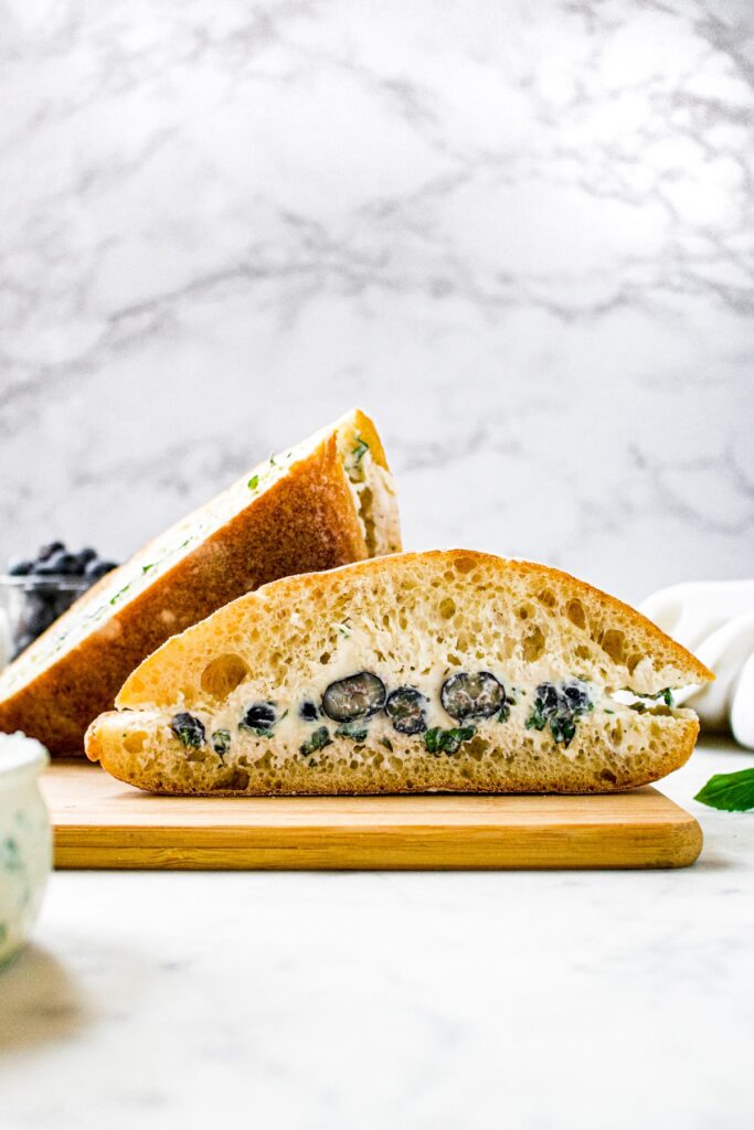 Head on shot of a vegan goat cheese and blueberry basil sandwich on a ciabatta roll sliced in half and sitting on a cutting board.