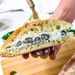 A hand holding half a ciabatta sandwich with creamy vegan goat cheese, fresh blueberries, and minced basil.