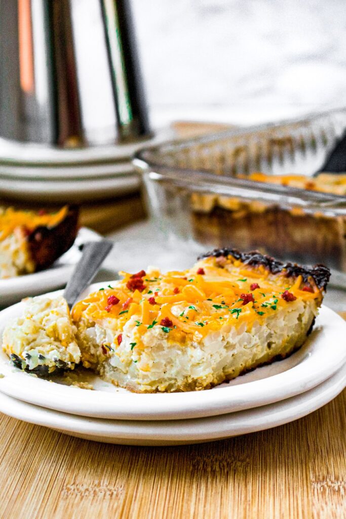 Head on shot of a slice of vegan hash brown casserole with a fork next to it that has a bite taken out of the casserole on it. There's another slice of casserole as well as the full baking dish in the background.
