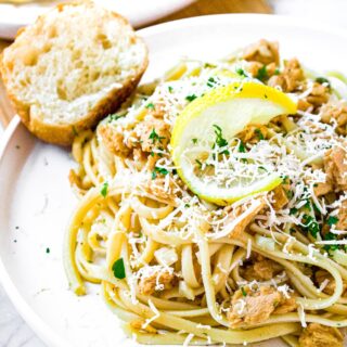 Close up photo of a pile of linguine in dairy free white wine sauce on a round white plate with a twisted lemon rind, minced herbs, and grated vegan parmesan on top