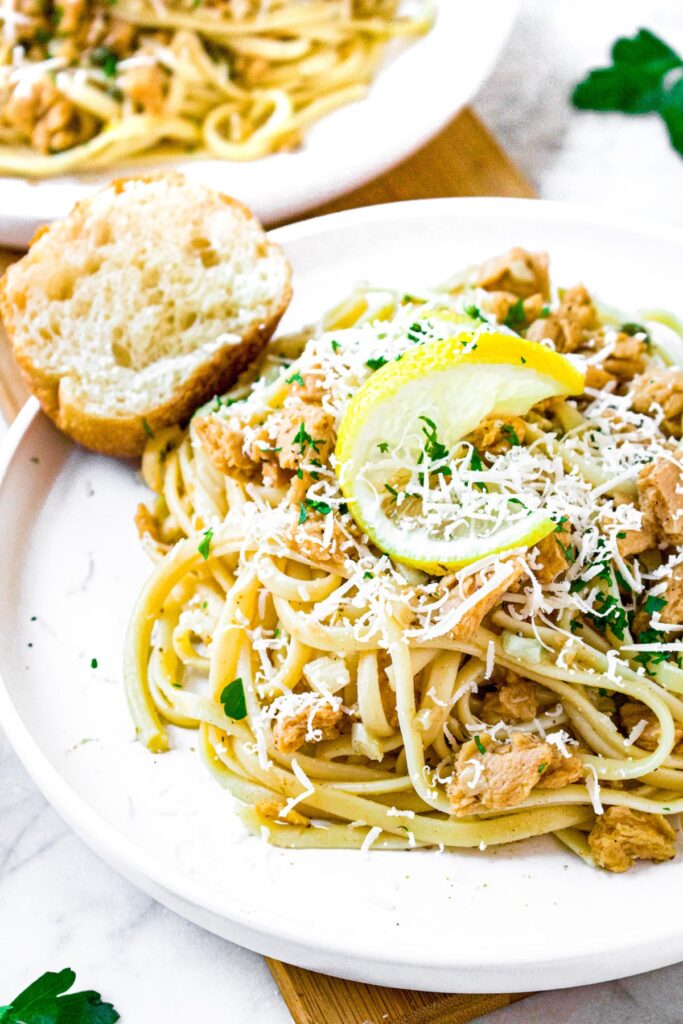 Close up shot of a pile of linguine in dairy free white wine sauce on a round white plate with a twisted lemon rind, minced herbs, and grated vegan parmesan on top
