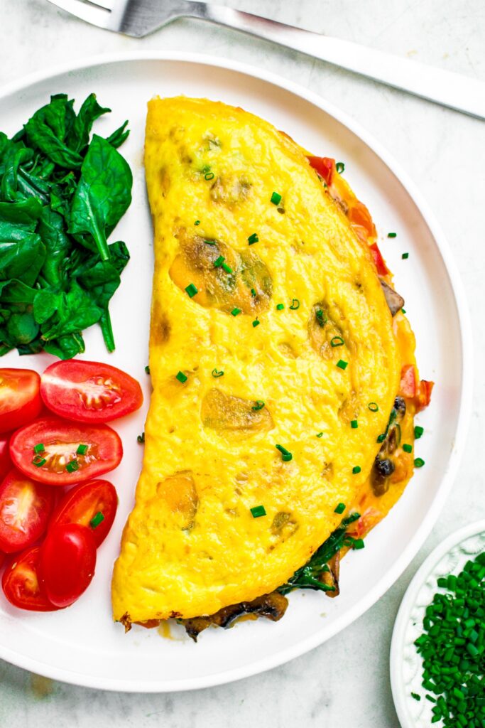 Close up overhead photo of a folded vegan omelet on a round white plate with spinach, mushroom, and dairy-free cheese filling spilling out the side