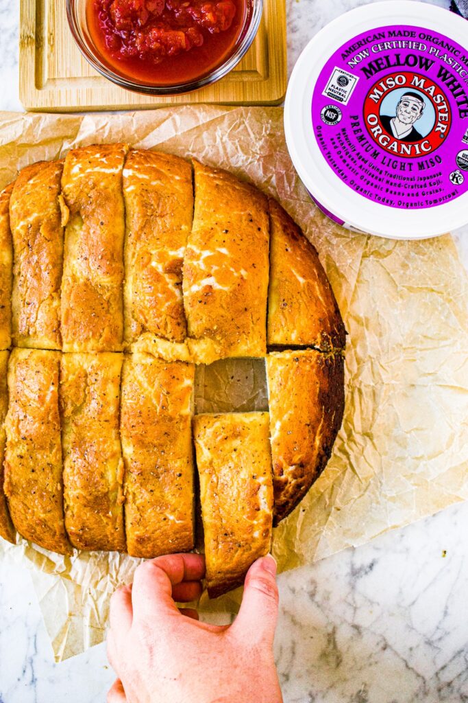 Overhead shot of sliced vegan cheesy bread with a hand pulling one slice out and a tub of Miso Master Organic Mellow White Miso Paste in the top right corner.