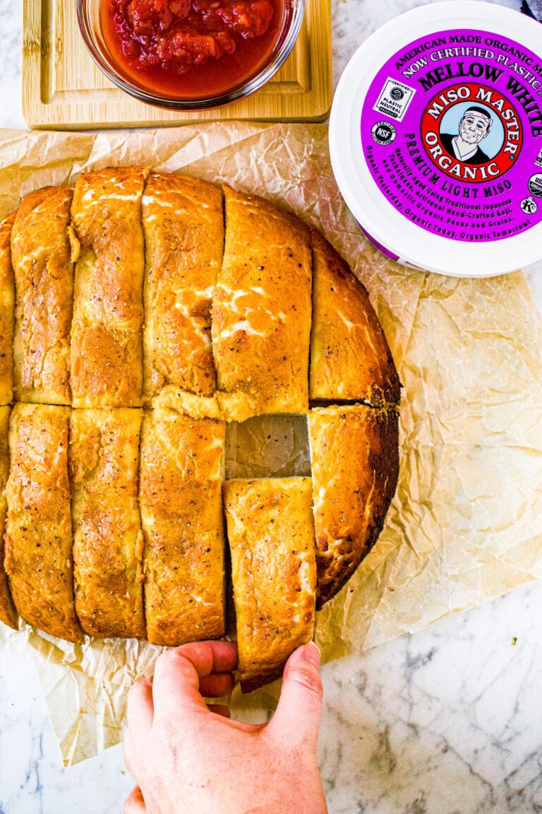 Overhead shot of sliced vegan cheesy bread with a hand pulling one slice out and a tub of Miso Master Organic Mellow White Miso Paste in the top right corner.